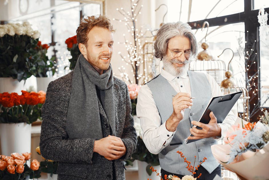 A florist with gray hair, beard, and glasses is holding a tablet while likely taking an order or reviewing floral arrangements inside a flower shop. Next to him, a young man with curly light brown hai