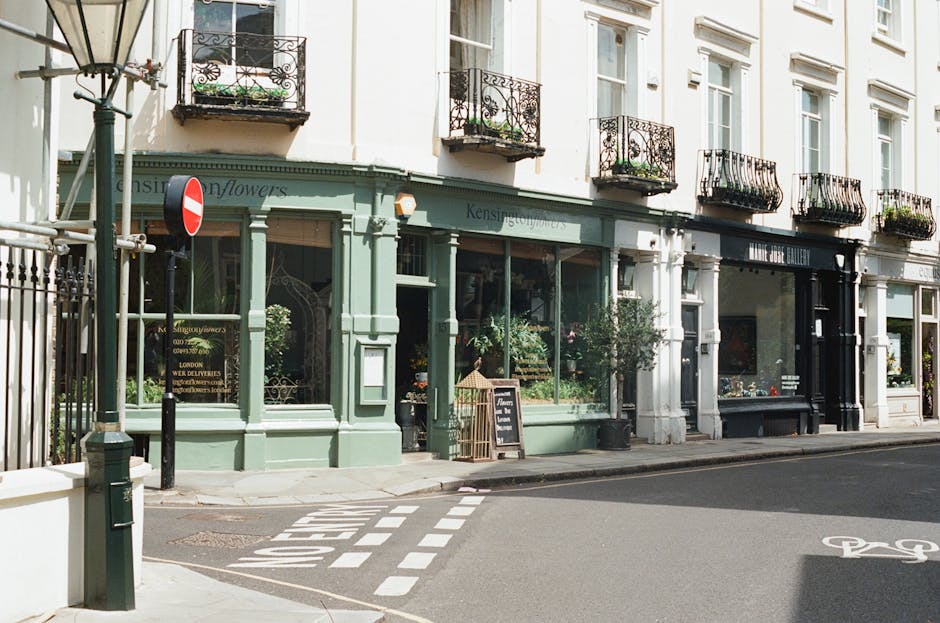 A street view of a florist shop with a pale green exterior, large display windows showcasing various flower arrangements, and a small chalkboard sign outside advertising flower arrangements. The shop