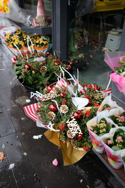 A festive floral arrangement featuring a variety of seasonal flowers and foliage, including pinecones, red berries, and small red and gold baubles, all incorporated into a lush bouquet with green pine