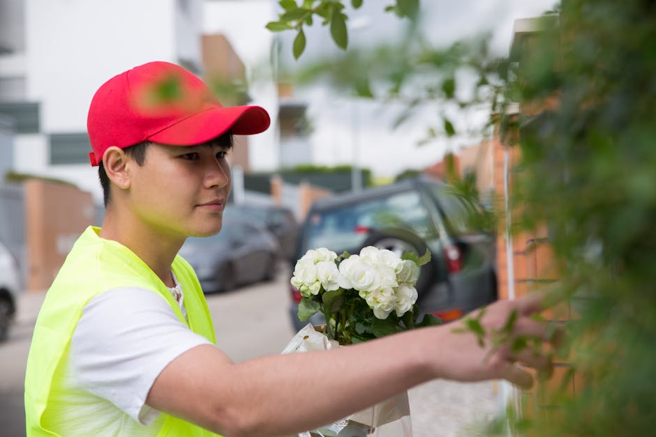 A delivery person wearing a red cap and a yellow high-visibility vest is holding a bouquet of fresh white roses with lush green leaves, wrapped in white tissue paper. They are outdoors near a resident