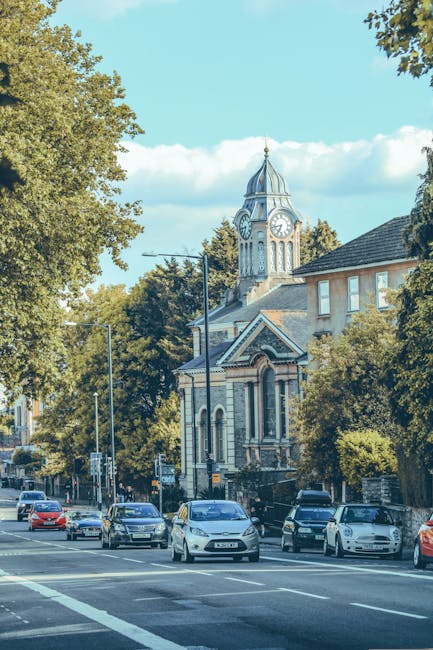 The image depicts a busy street scene in Rayners Lane during daytime, with multiple cars parked along the roadside and some in motion. Tall, leafy trees line the street, their green foliage partially 
