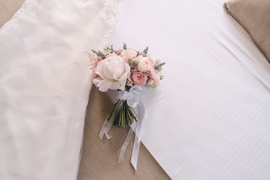 A delicate bouquet of fresh flowers resting on a beige upholstered surface. The arrangement features large white peonies, soft pink roses, and smaller sprigs of lavender and muted greenery, all expert