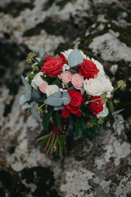 A bouquet featuring a combination of red roses, pale pink ranunculus, and white peonies, accented with grey-green eucalyptus leaves and other foliage. The flowers are arranged in a compact, rounded st