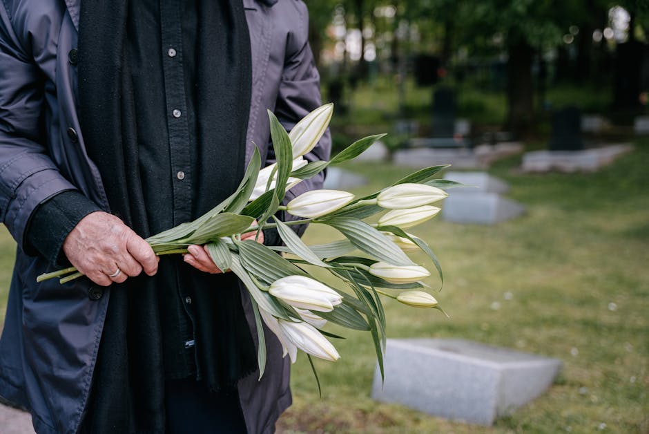 A person wearing a dark grey jacket and black shirt holding a bouquet of fresh white lilies with green leaves, arranged in a natural style. The bouquet features long-stemmed lilies with closed buds an