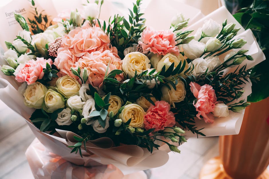 A floral bouquet featuring soft pink carnations, creamy white roses, and white lisianthus, complemented by dark green foliage and eucalyptus leaves. The flowers are arranged in a loosely gathered styl