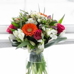 Designed to reflect the welcoming and artistic spirit of Rayners Lane, this luxurious bouquet is a showcase of both colour and craftsmanship. Creamy and soft peach gerberas form a gentle backdrop while vivid orange counterparts spark energy and excitement, mirroring the vibrant social life that thrives around Rayners Lane station. Plush magenta roses nestle between the gerberas, their bold tones echoing the notes of celebration that fill local homes during festive times. Pristine white alstroemeria and clusters of petite filler blooms add brightness and a frothy texture, enveloping each blossom in light, much like the community's signature blend of calm and vivacity. The arrangement is cradled by arching, broad aspidistra leaves, whose deep emerald tones offer contrast and structure, reminiscent of the leafy lanes that wind through West Harrow. Finer green leaves and sprigs weave between the larger flowers, infusing the ensemble with a layered sense of abundance. Through the transparent vase, every stem is visible, underscoring the garden-fresh quality. The bouquet, illuminated by soft, even daylight, is a symphony of optimism, making it a fitting centerpiece for celebrations, brunches, or as a token of appreciation. Each element, chosen by the florists at Flowers Rayners Lane, captures the area's unique blend of tradition and modern style.